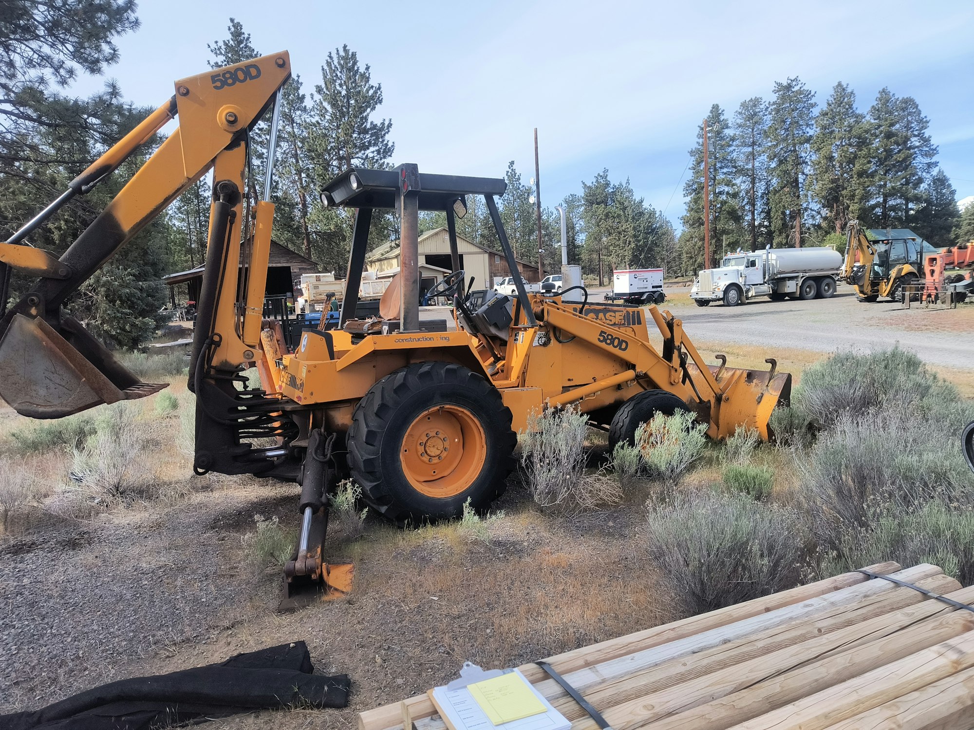 A yellow Case 580D backhoe loader parked on a gravel area, surrounded by trees and other construction vehicles in the background.