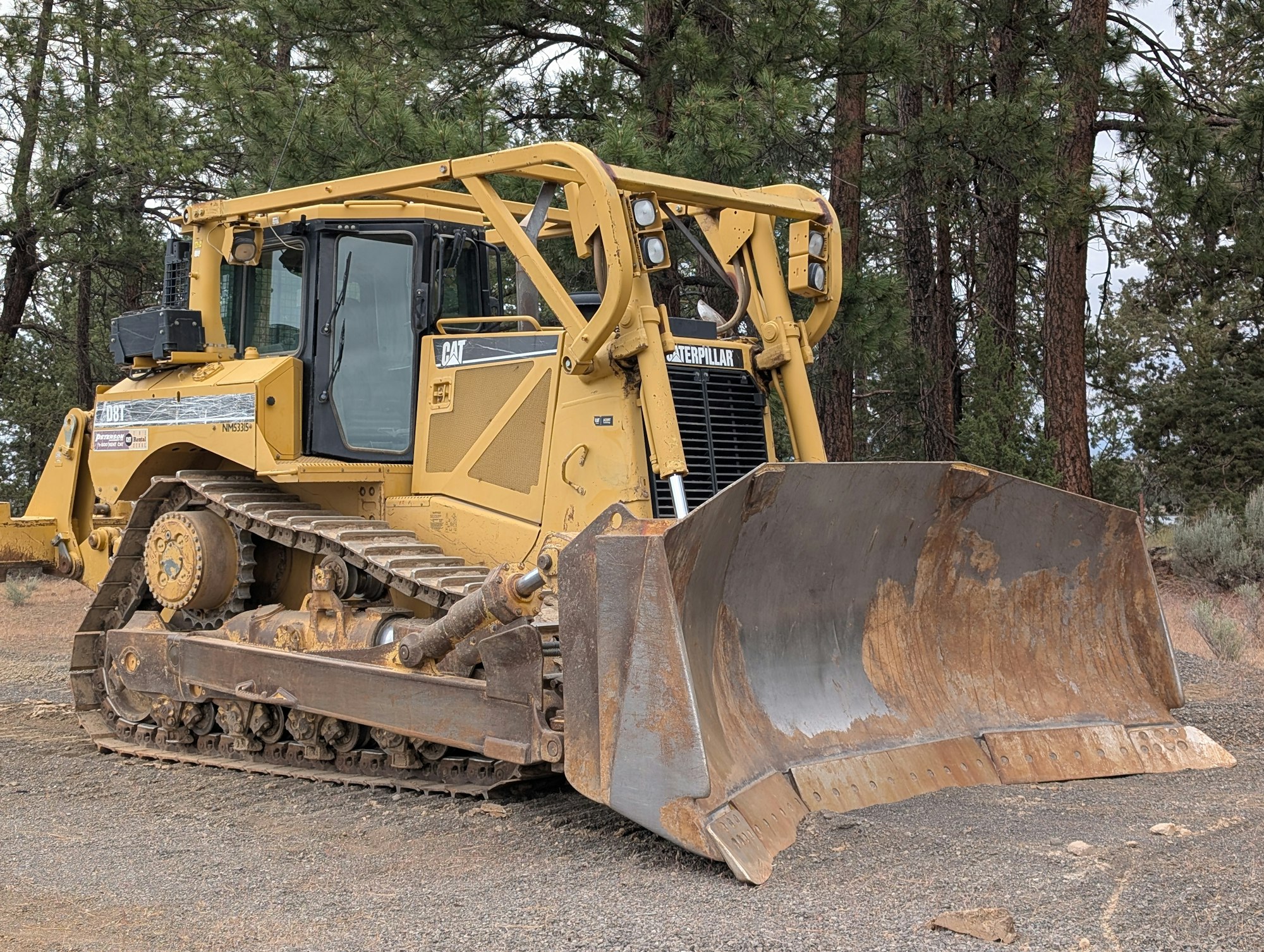 A yellow bulldozer parked on gravel, surrounded by trees, with a large metal blade at the front.