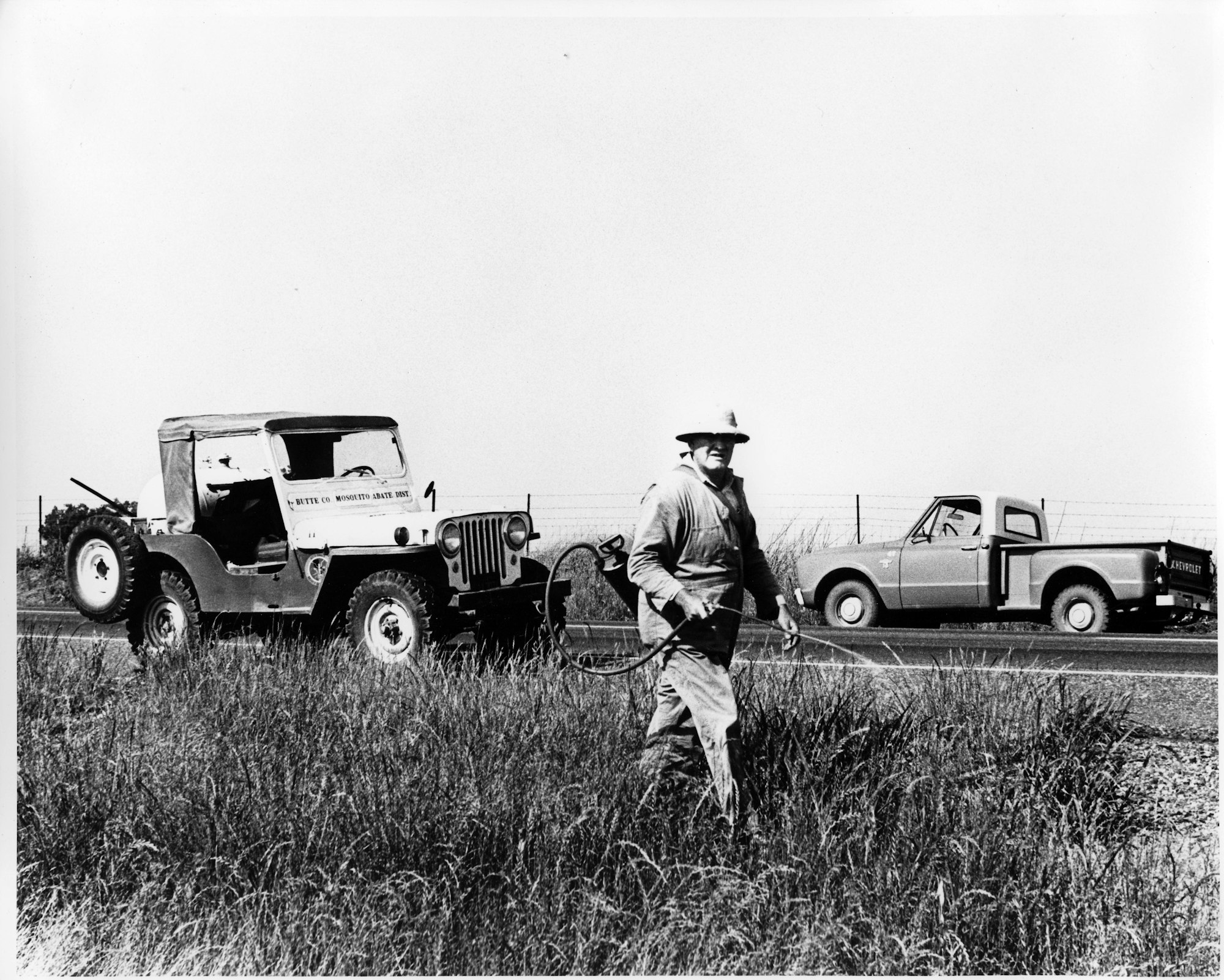 A person sprays weeds by the roadside. There's a jeep and a pickup truck in the background.