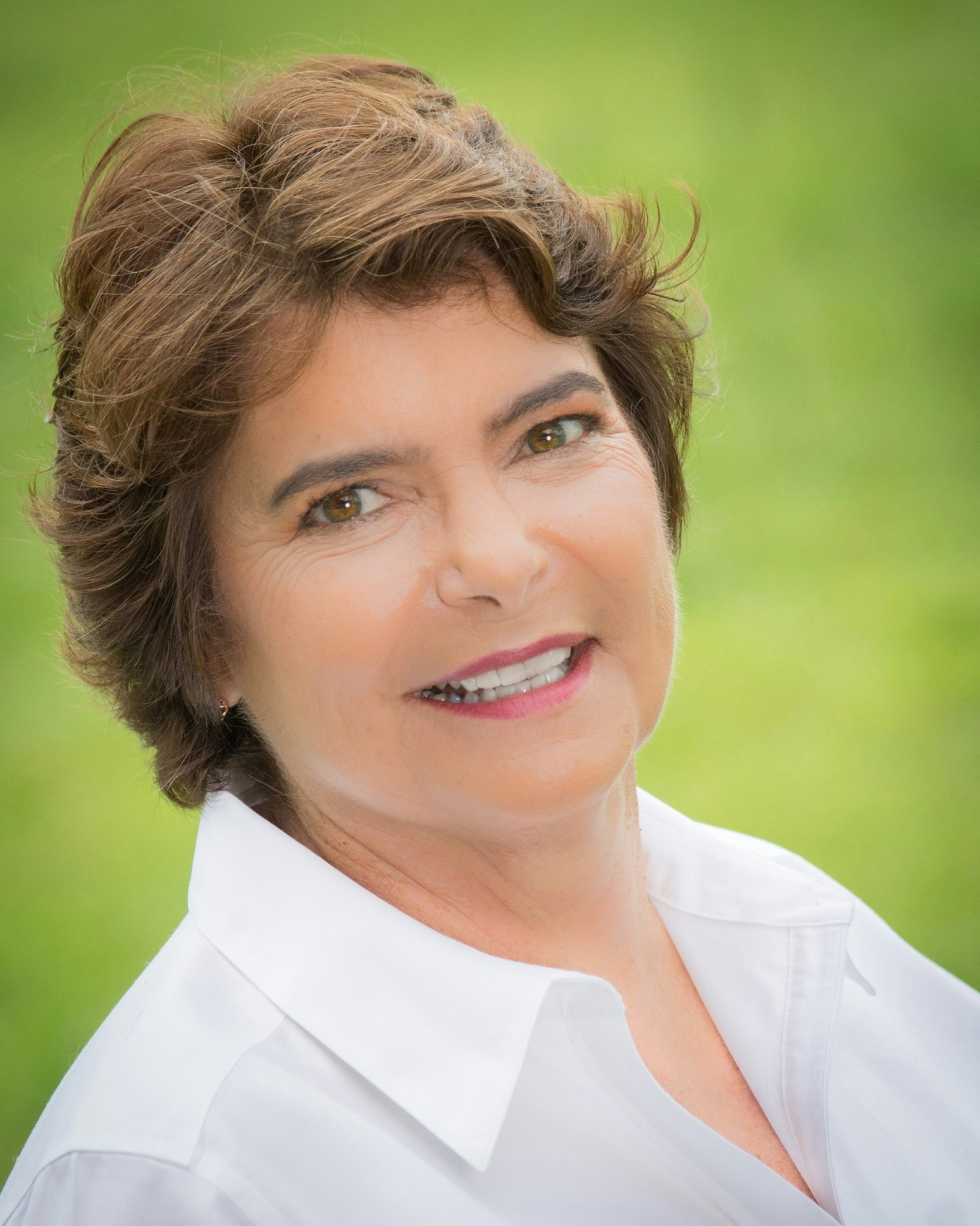 A portrait of a smiling woman with brown hair, wearing a white shirt, with a blurred green background.