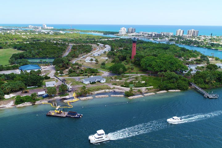 waterway with dredge and boats navigating by in front of Jupiter Lighthouse with nearshore breakwaters being constructed