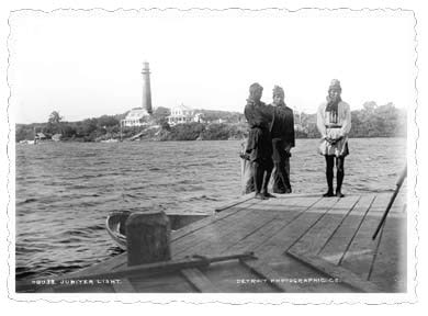 1891 black and white photo with three people standing on dock with water and the Jupiter lighthouse in the background