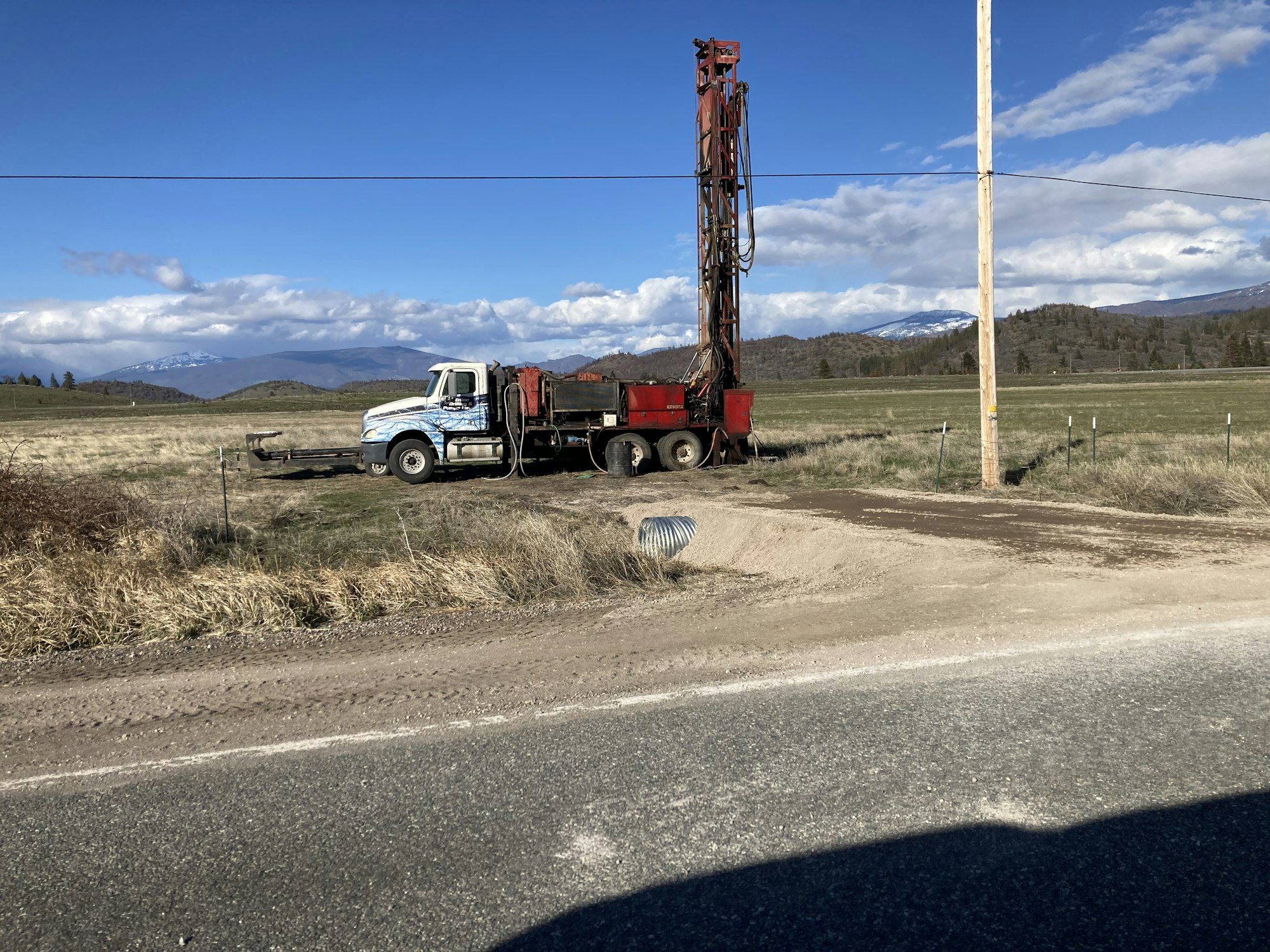 A truck with drilling rig equipment by a dirt road, grassy field, distant mountains, and a clear sky.