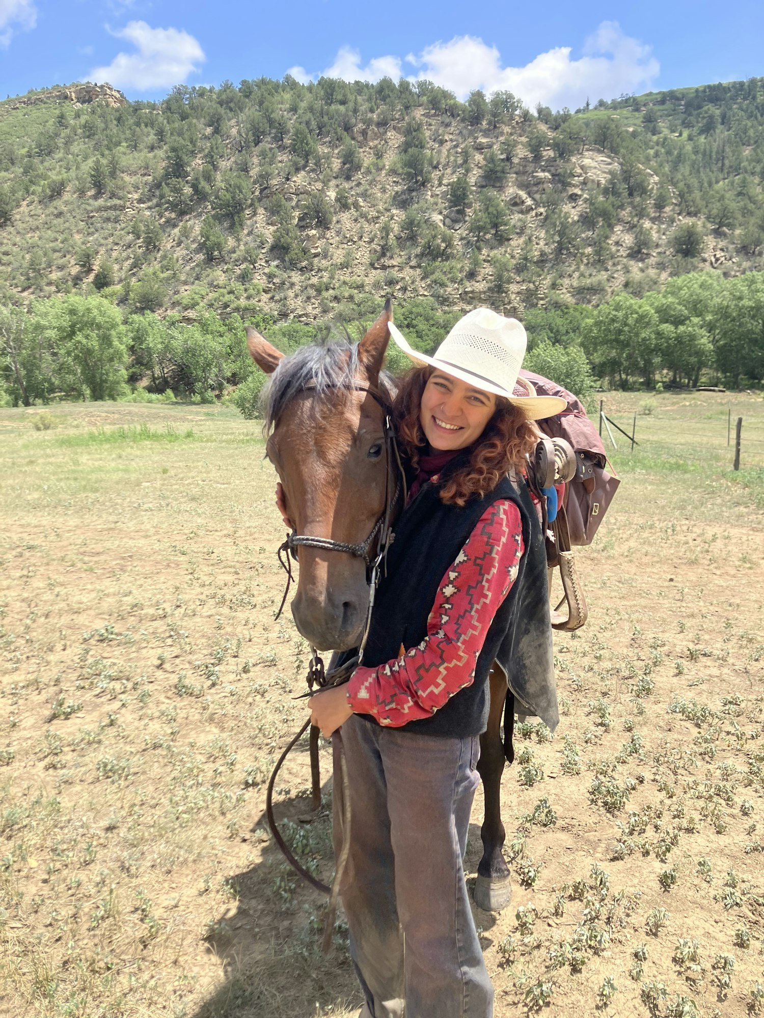 A smiling person in a cowboy hat with a horse, set in a scenic outdoor landscape with hills and trees.
