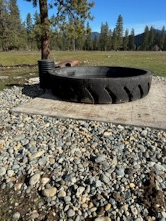 The image shows a large, round tire used as a fire pit, surrounded by gravel on a grassy area with trees in the background.