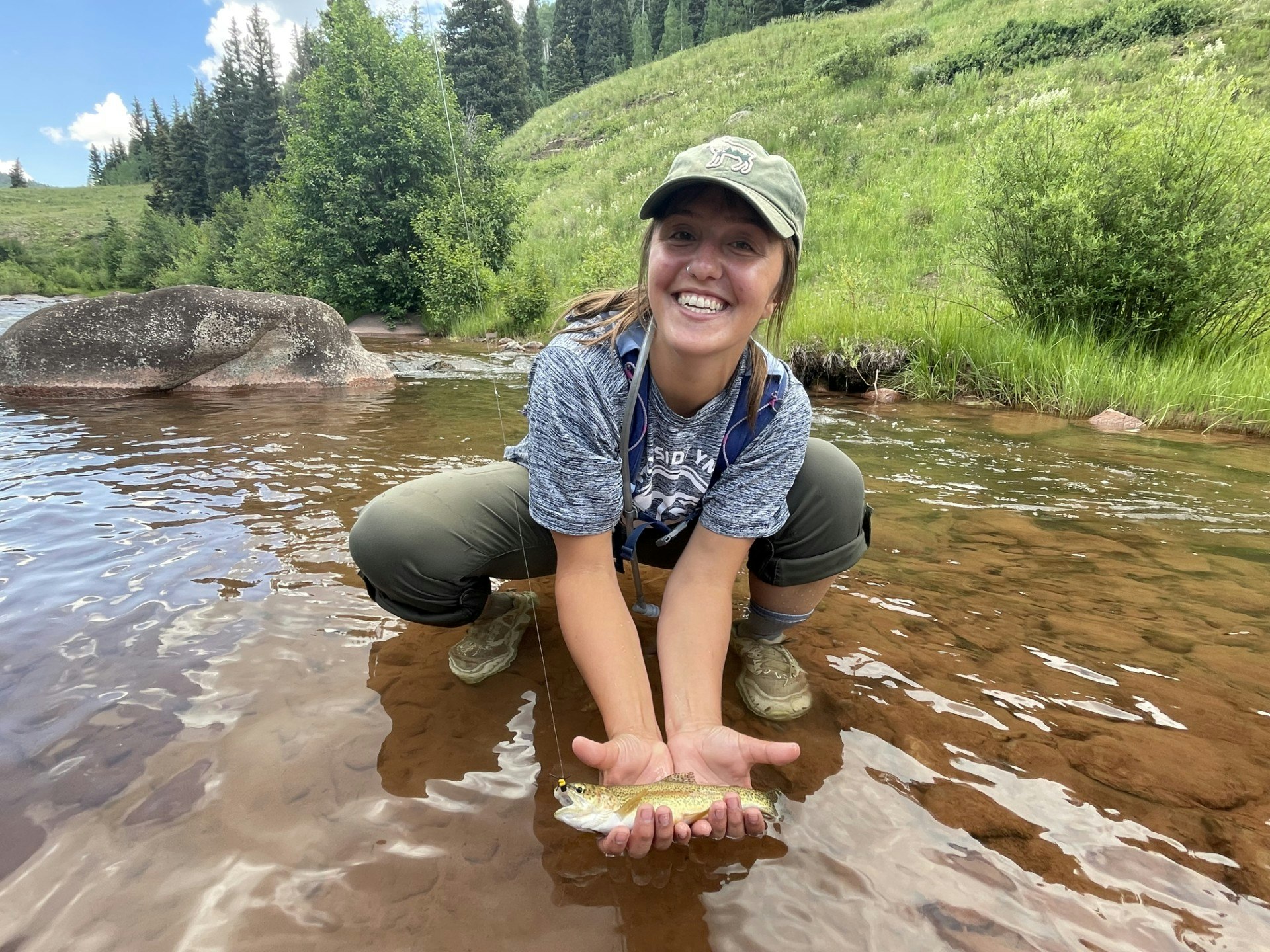 A smiling person crouched by a stream, holding a small fish, with greenery and a fishing rod in the background.