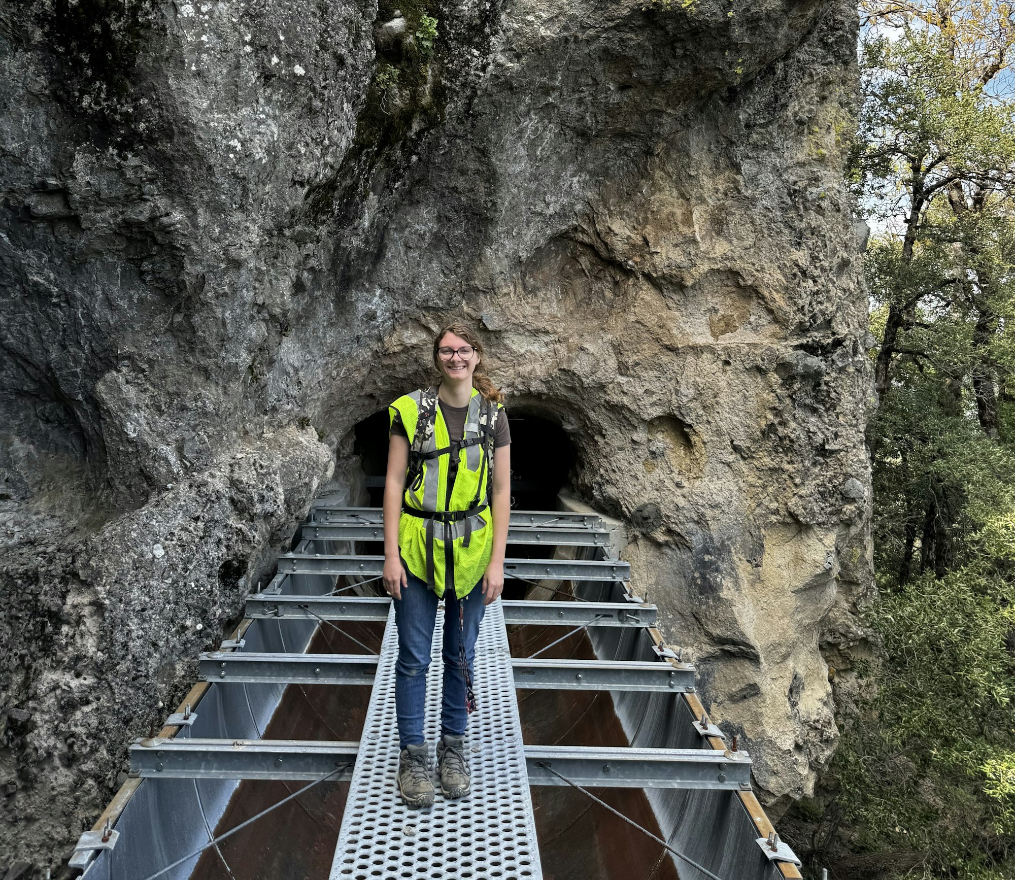 A person in a high-vis vest stands on a metal walkway by a rocky cave entrance, surrounded by greenery.