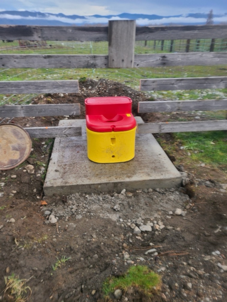 A bright red and yellow container sits on a concrete slab, surrounded by a rustic wooden fence in a grassy area.