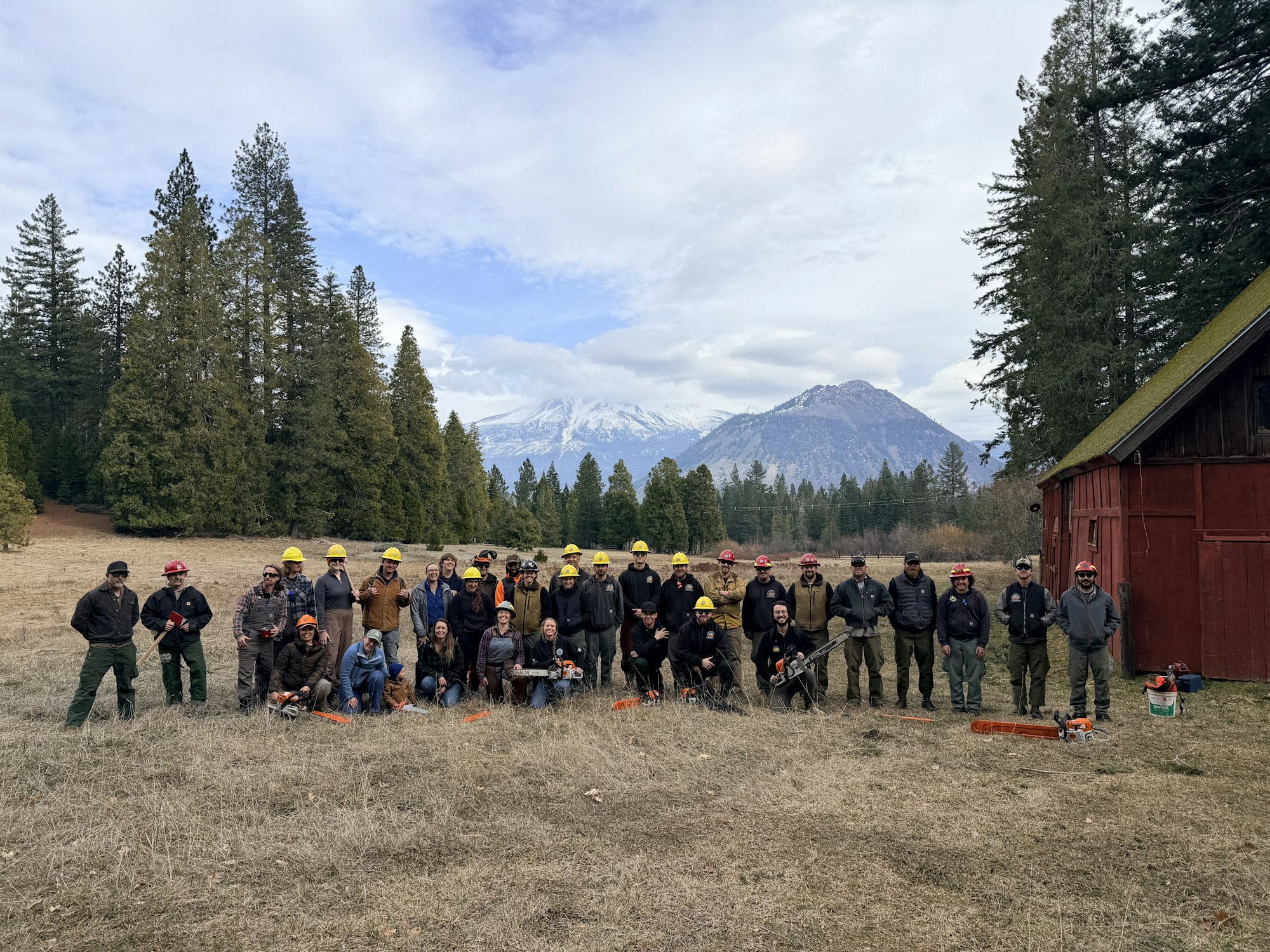 A large group of people in safety gear pose for a photo outdoors, surrounded by trees and mountains, with tools in hand.