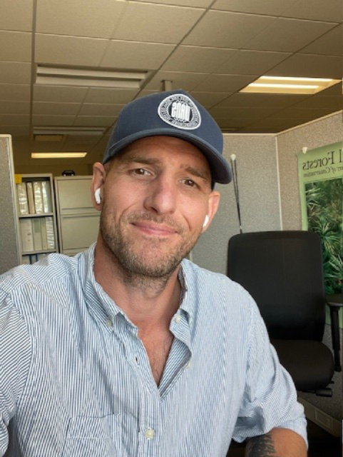 A person wearing a cap and earbuds is in an office setting, smiling at the camera.