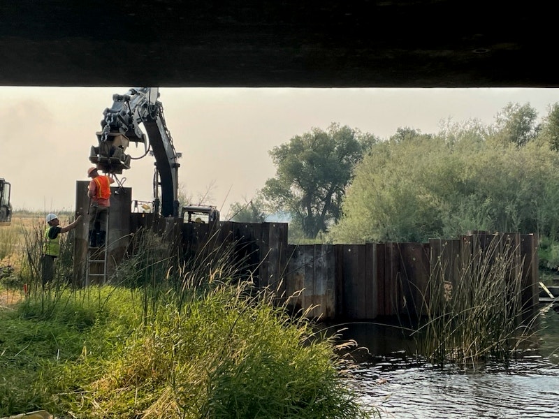 A construction site near water, featuring workers, machinery, and metal barriers surrounded by greenery.