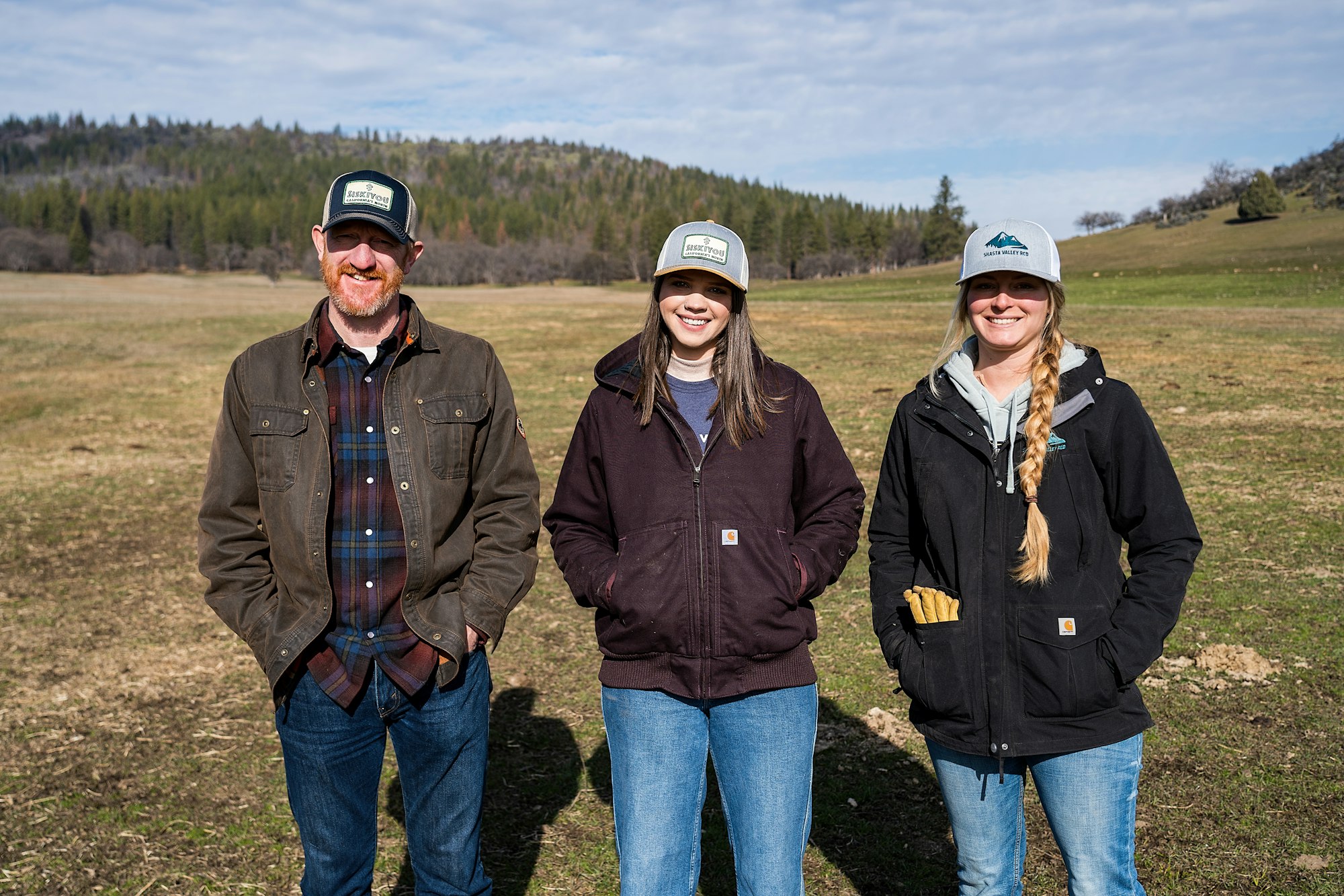 Three people standing in a field with a forest in the background, all wearing casual clothing and caps.