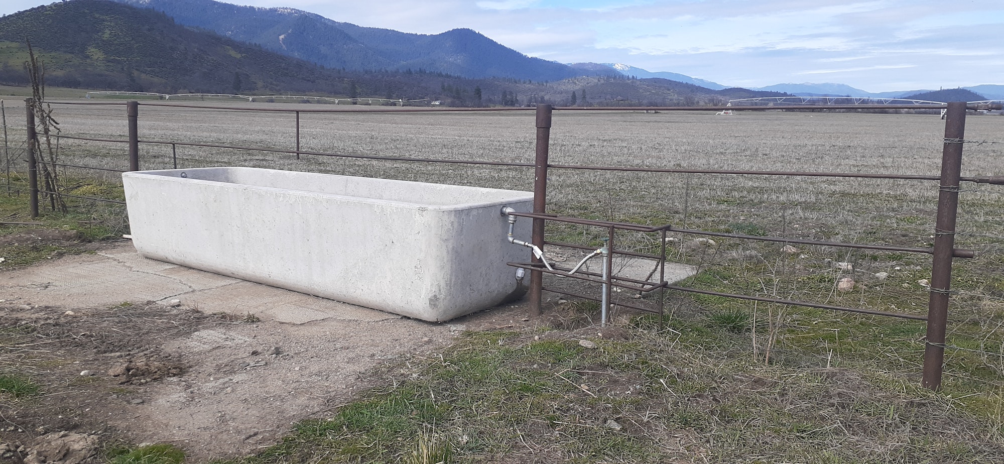 A concrete trough next to a fence, set in an open field with mountains in the background.