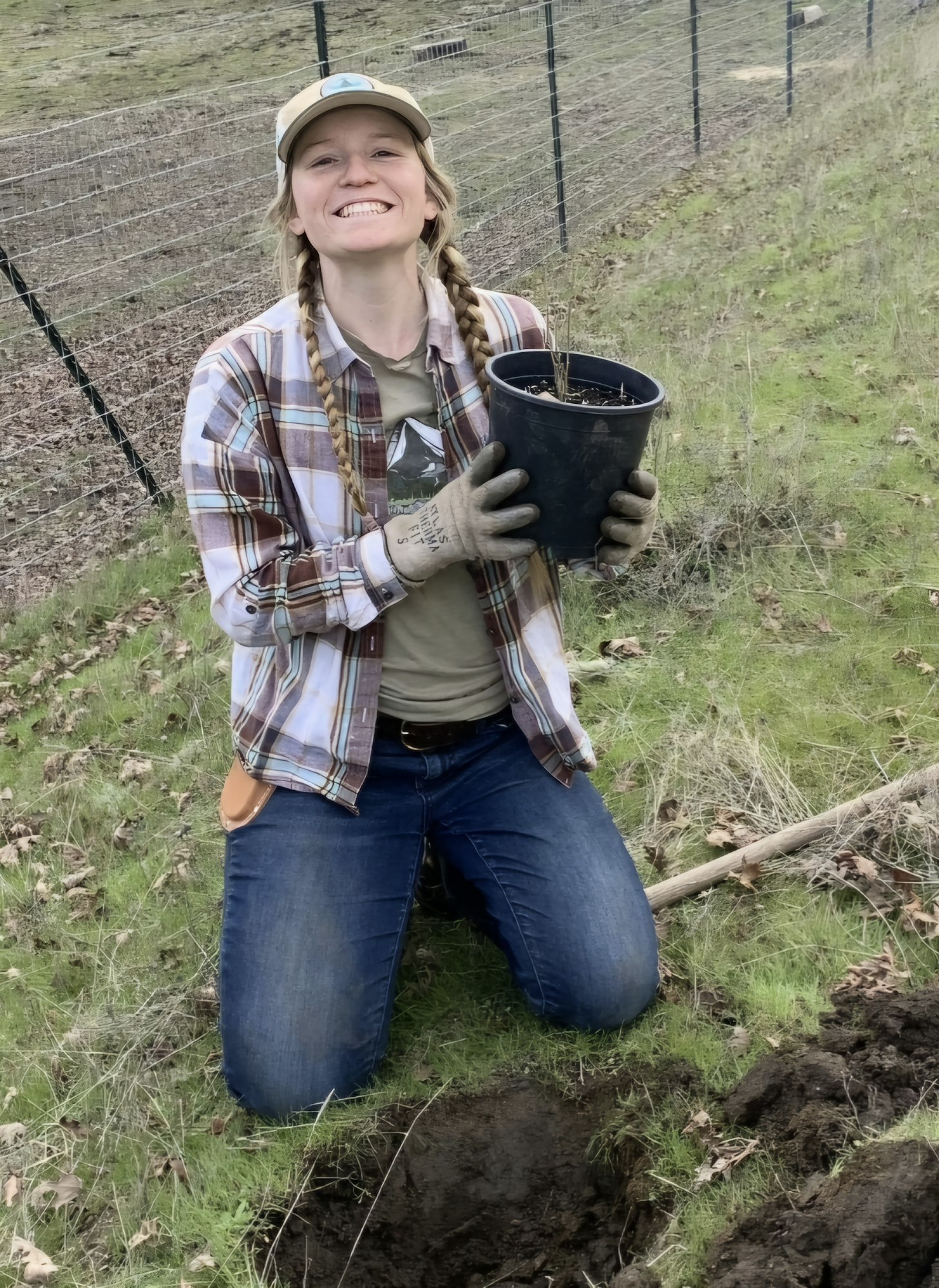 Person kneeling outdoors, holding a plant in a pot, wearing gloves and a plaid shirt. Smiling with a grassy background.