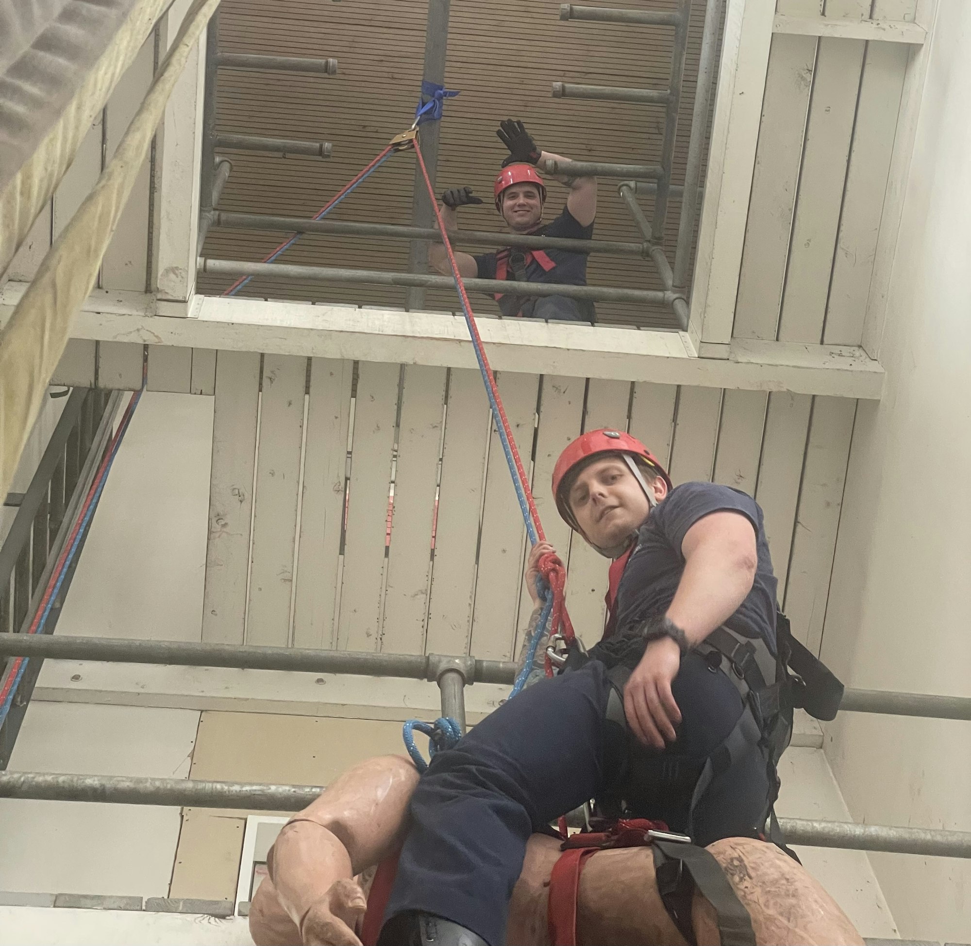 Two individuals in helmets are using ropes in a simulated rescue scenario, one climbing while the other assists from above.
