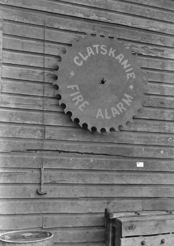 An old circular saw blade repurposed as a sign for "CLATSKANIE FIRE ALARM" on a wooden wall.