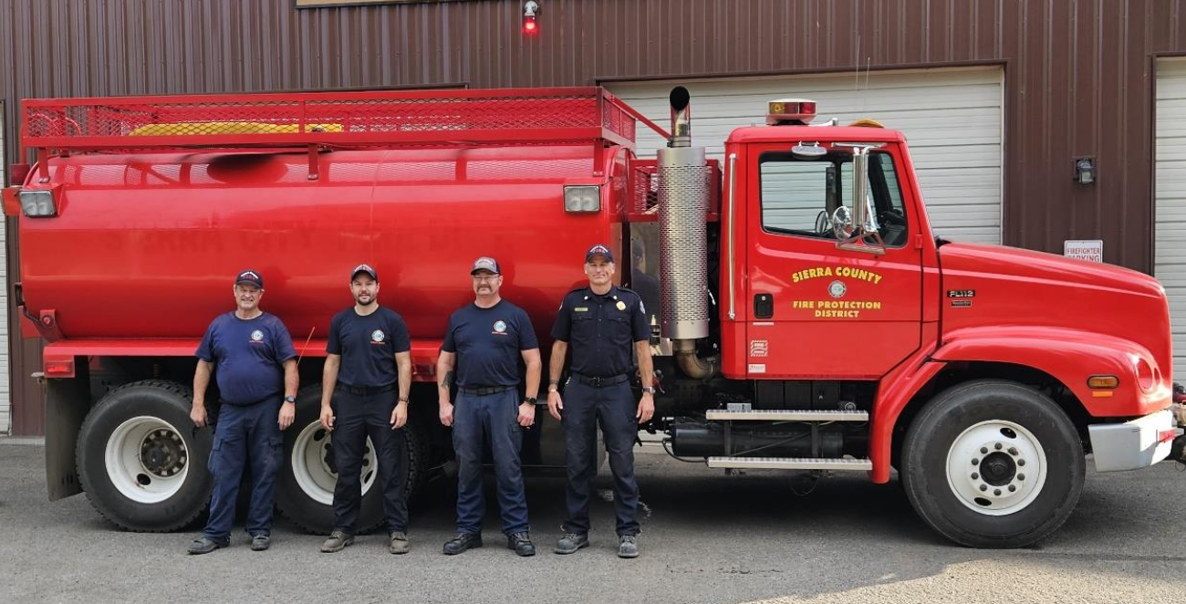 Four firefighters standing in front of a red fire truck labeled "Sierra County Fire Protection District."
