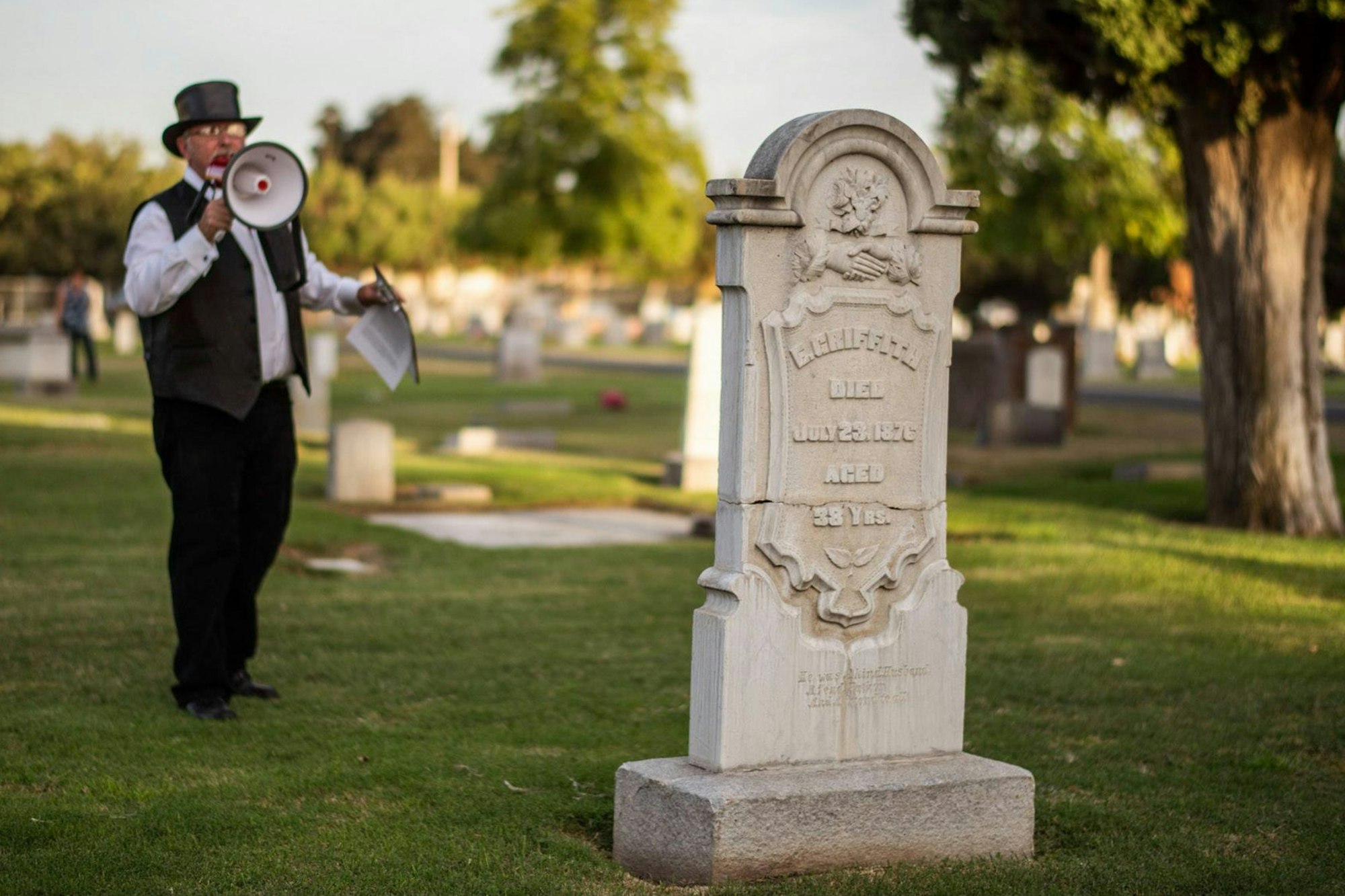 Man with megaphone near tombstone in cemetery, wearing a top hat and vest, holding papers.