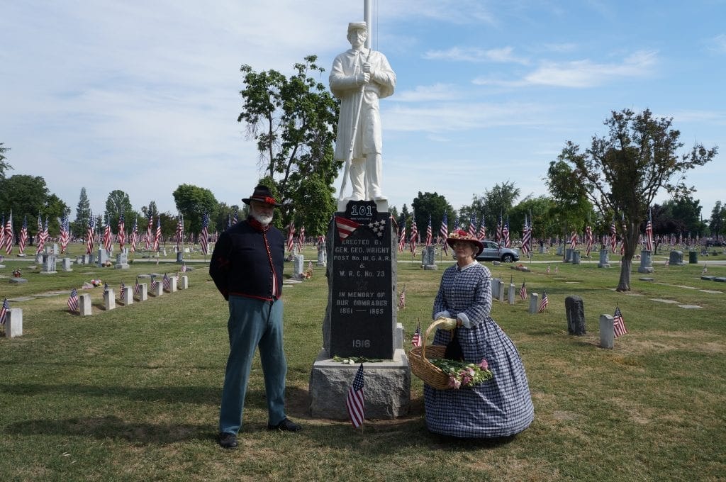 Two people in historical attire beside a memorial with flags in a cemetery.