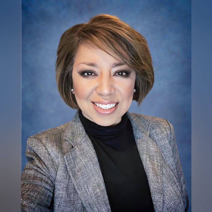 A smiling person with short hair, wearing a blazer and a turtleneck, against a blue background.