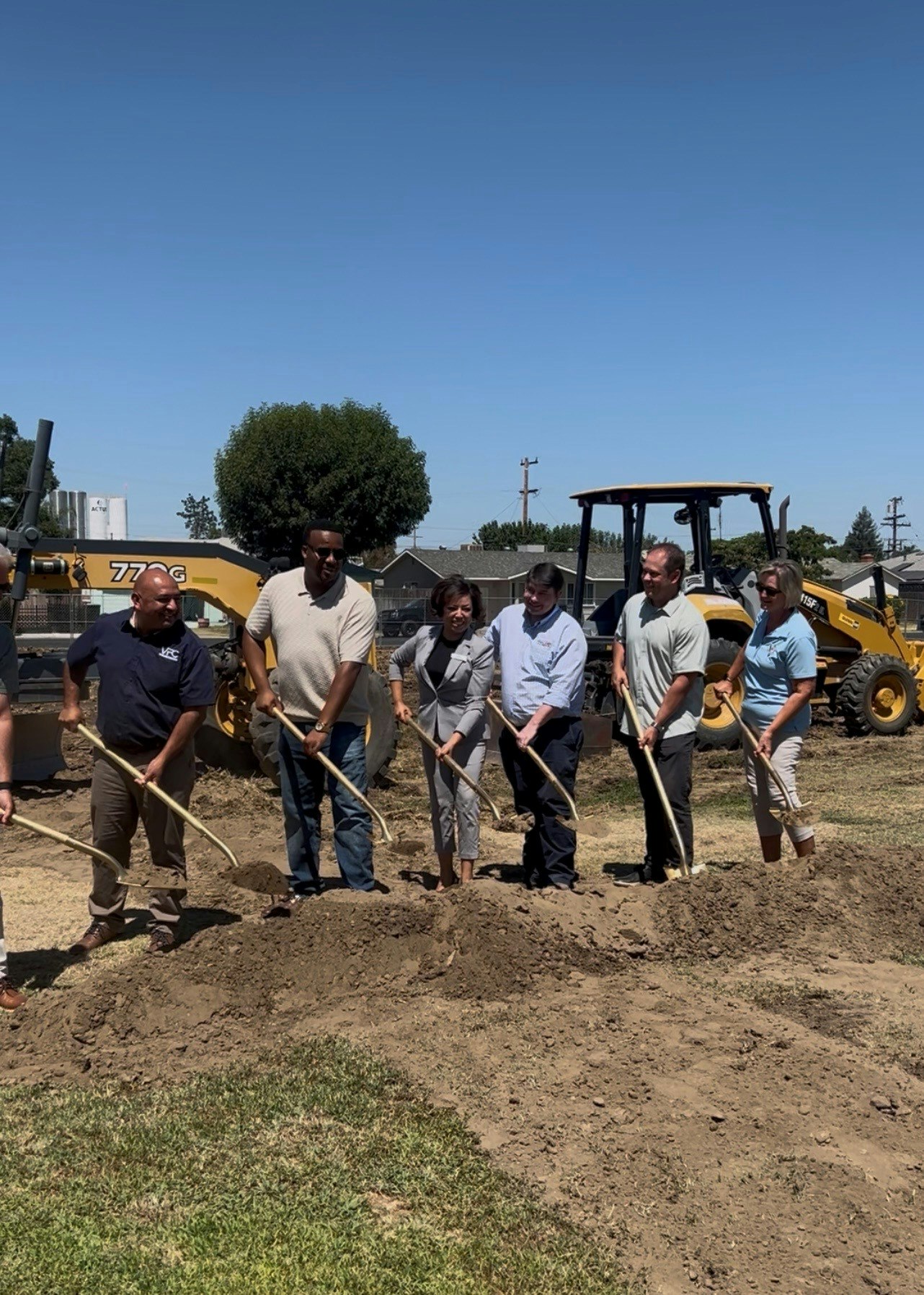 A group of people is participating in a groundbreaking ceremony with shovels, set against a clear sky and construction equipment.