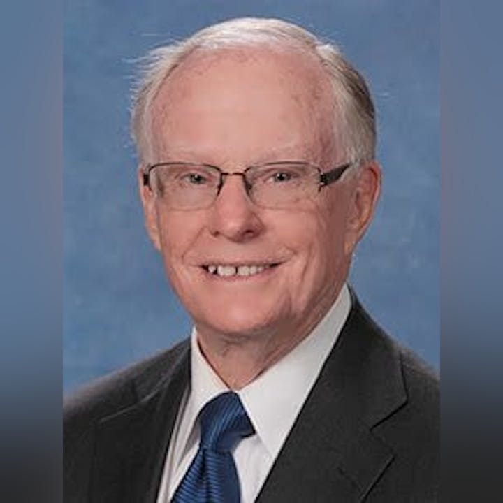 A portrait of an elderly man with glasses, wearing a suit and tie against a blue background.