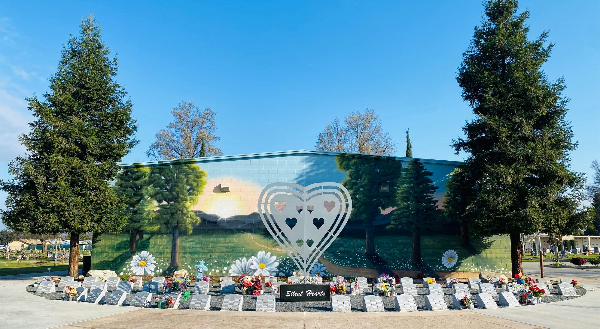 A mural with trees and hearts, flanked by real trees, with memorial stones and flowers in front.