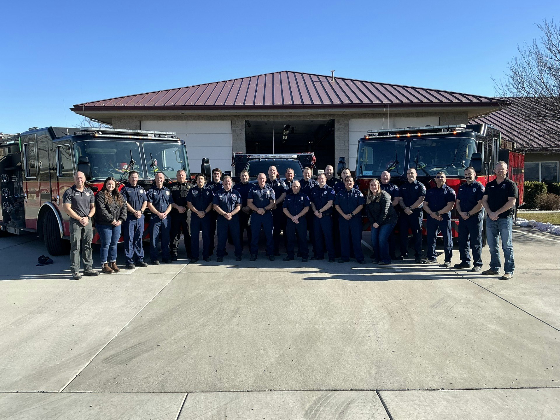 A group of people, possibly firefighters, pose in front of two fire trucks at a fire station.