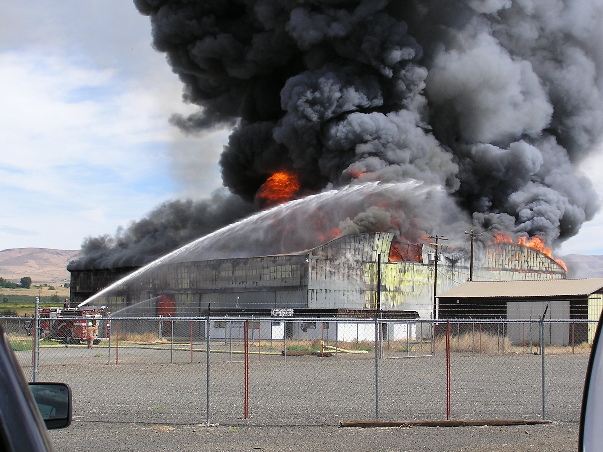 A large industrial building on fire with firefighters battling the blaze, thick black smoke billowing into the sky.