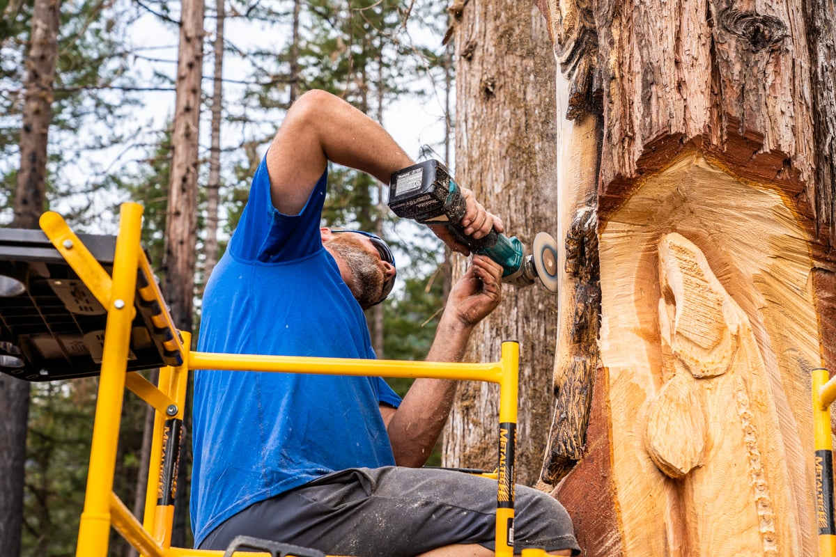 A person carving a tree sculpture with a power tool, standing on a lift.