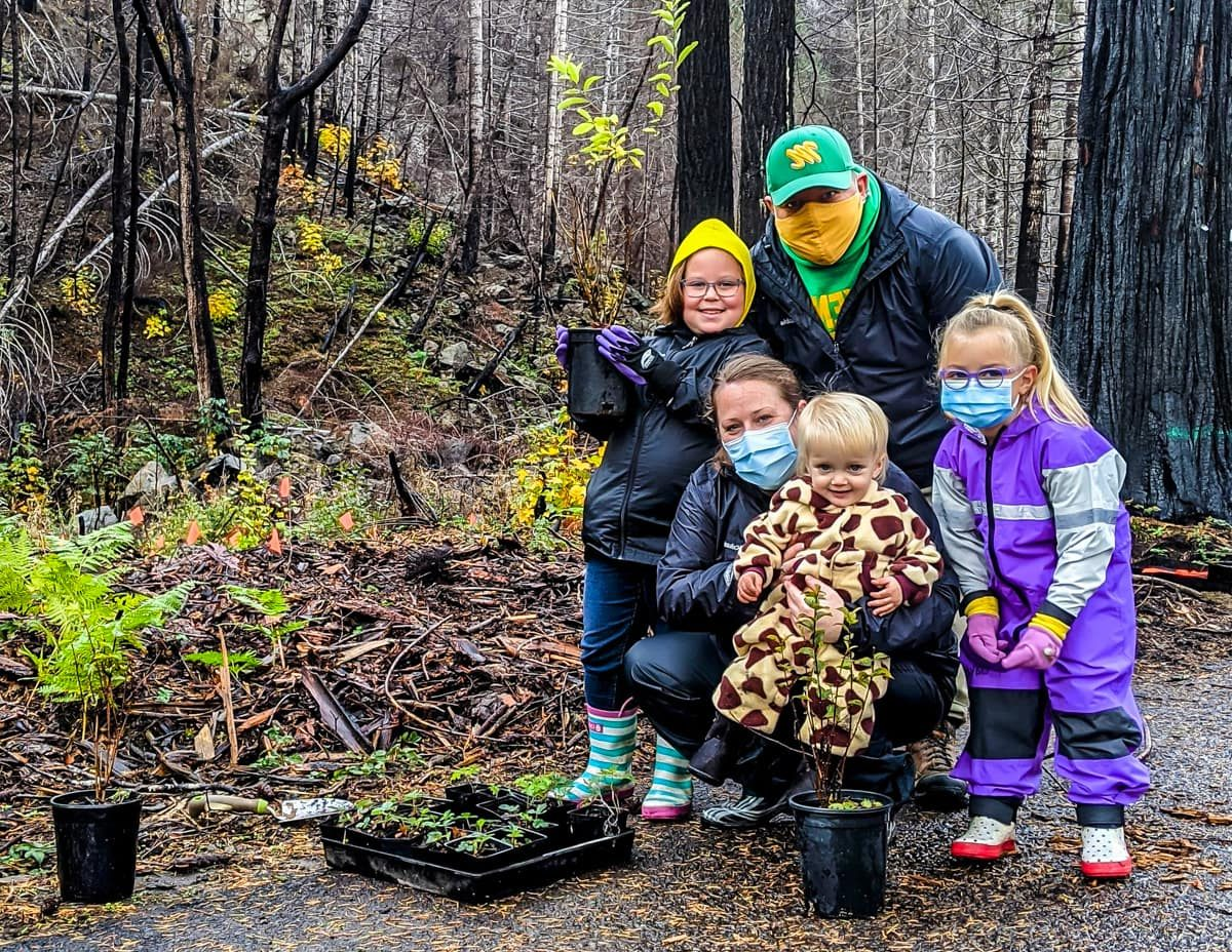 A family with young children, wearing masks, standing in a forest with saplings and gardening tools, suggesting a tree-planting activity.