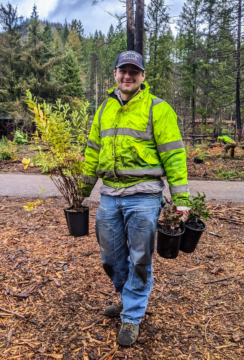 A person in a high-visibility jacket holding two potted plants, standing in a forested area with others in the background.