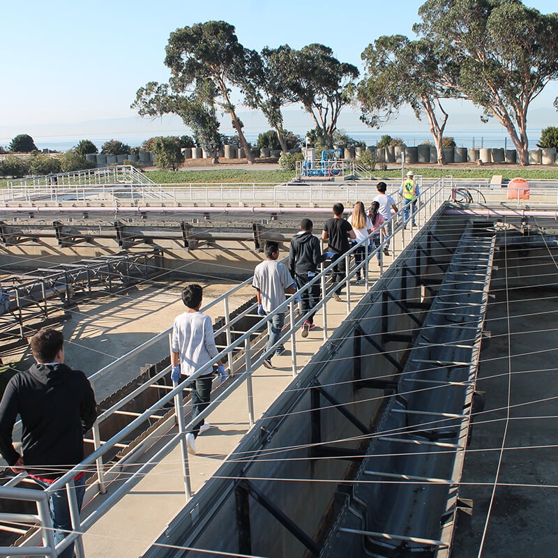 A group of people walking on a walkway near industrial tanks surrounded by trees and open landscape.