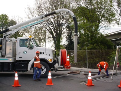 A vacuum truck and workers in orange vests operate near a manhole, surrounded by traffic cones.
