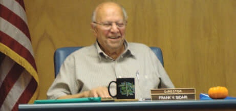 An elderly man sits at a desk with a nameplate, mug, orange object, and an American flag in the background.