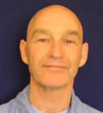 A person with a shaved head and wearing a blue shirt, set against a dark blue background.