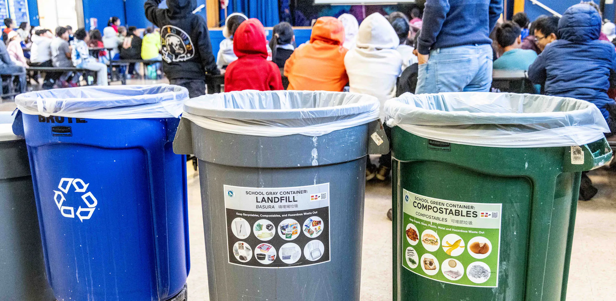 Three waste bins labeled for recycling (blue), landfill (gray), and compostables (green) are positioned near a seated audience.