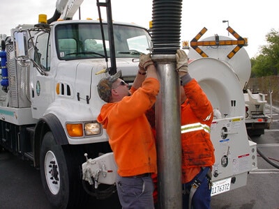 Two workers in orange safety gear operate a vacuum truck, handling a large hose or pipe in an urban setting.