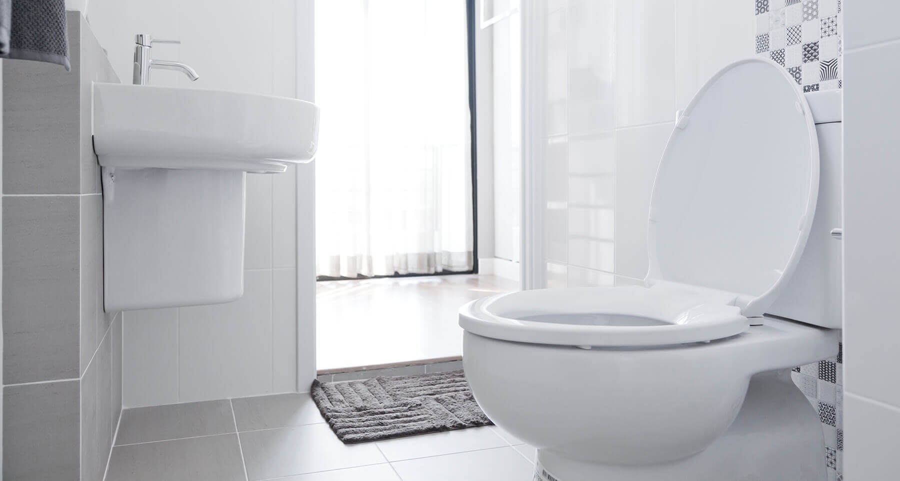 Modern bathroom with a white toilet, sink, and grey floor tiles; sunlight through sheer curtains.