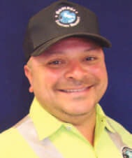 A person smiling, wearing a black cap with a logo and a high-visibility shirt in front of a blue background.
