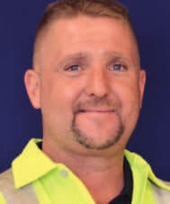 A person wearing a yellow collared shirt, smiling in front of a dark blue background.