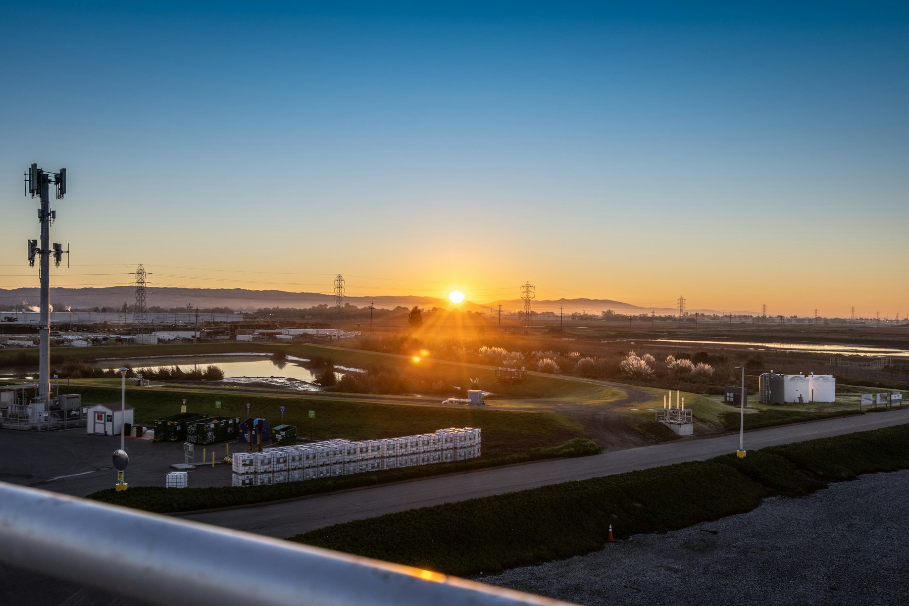 A sunrise over a scenic landscape with a waterway, green fields, utility poles, and industrial structures.