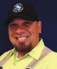 A smiling person wearing a black cap and a bright green shirt with a dark blue background.