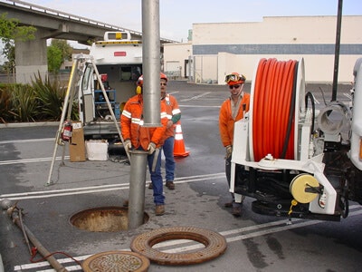 Workers in orange gear, inspecting or cleaning a sewer, with equipment and an open manhole nearby.