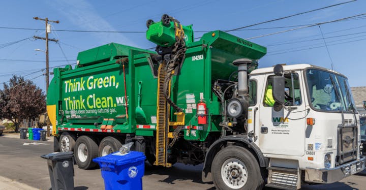A green garbage truck with "Think Green, Think Clean" slogan is collecting waste from recycling bins on a residential street.