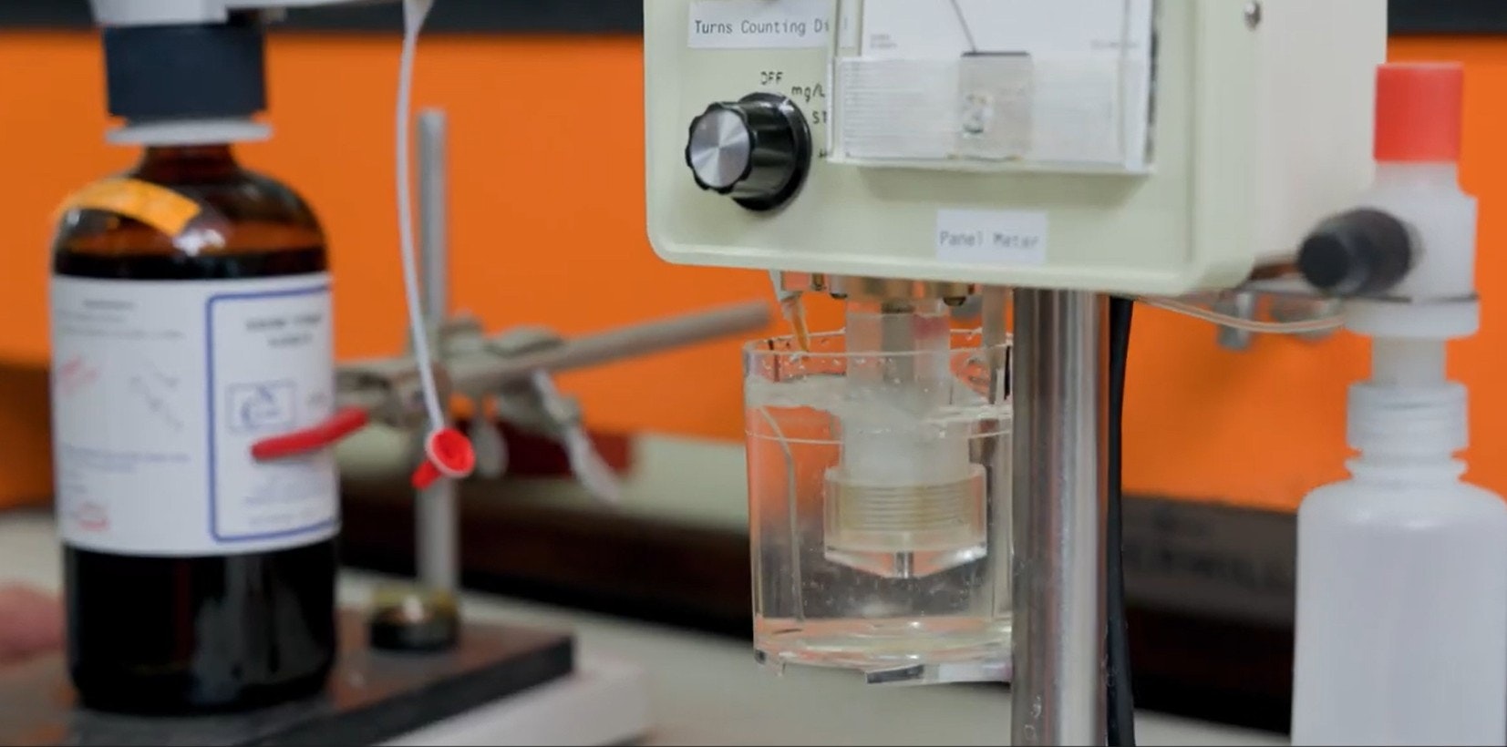 The image shows laboratory equipment, including a container with liquid and a digital meter for measuring parameters.