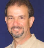 A man with a goatee, wearing a beige shirt, smiling against a dark blue background.