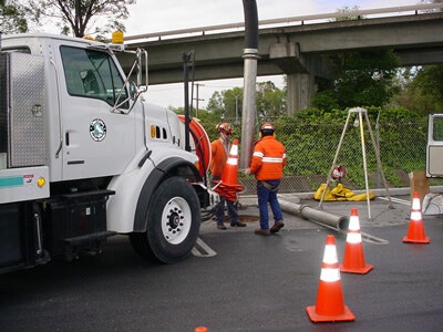 Workers in safety gear manage equipment next to a truck, surrounded by traffic cones, under an overpass.
