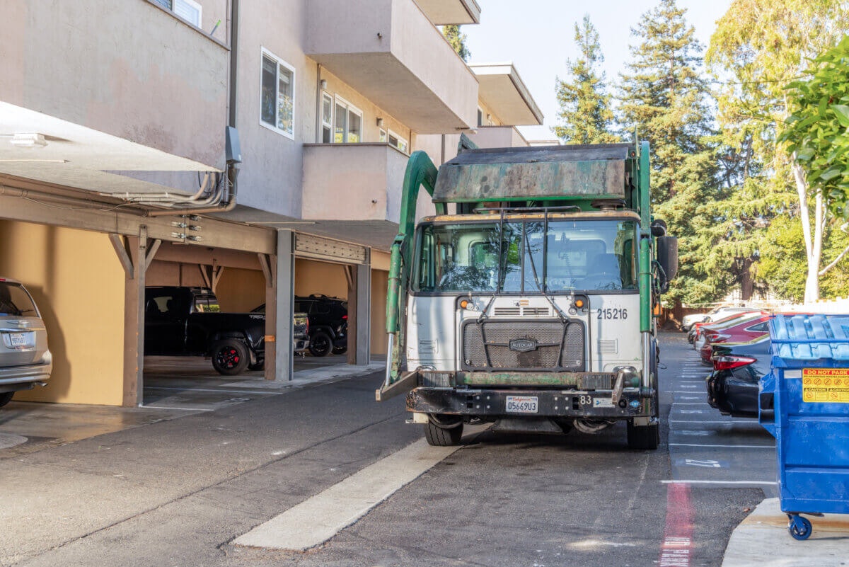 Garbage truck in a parking area near parked cars and apartments. Nearby is a blue dumpster.