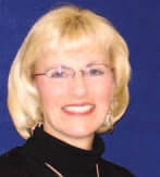 A person with short blonde hair and glasses smiling against a blue background.